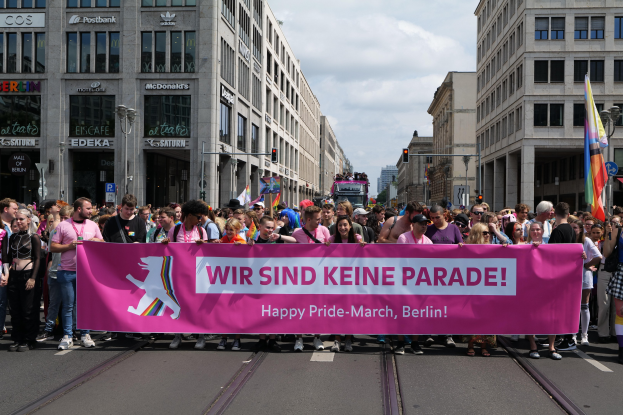 Gruppe von Menschen, die eine rosafarbene 'Happy Pride March'-Fahne auf einer Berliner Straße halten, mit Gebäuden, Laternen und Verkehrszeichen in der Straße unter einem bewölktem Himmel.