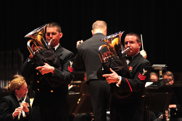 Eine Gruppe von Männern in Militäruniformen, die Euphoniums auf einer Bühne spielen, einige halten Stöcke und andere sitzen mit verschiedenen Instrumenten, vor einem dunklen Hintergrund.