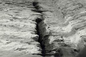 Schwarzes und weiße Foto eines großeen Risses im Boden, wahrscheinlich ein Entwässerungskanal, mit einem sichtbaren Wasserzeichen auf der linken Seite.