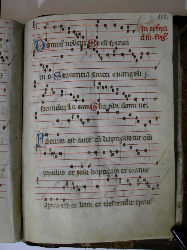 Ein aufgeschlagenes Buch mit musikalischen Noten auf seinen Seiten, identifiziert als "bibl sainte-geneviève, ms 0060, f 002v-002", liegt auf einer Oberfläche, mit sichtbarem Text auf der Seite.