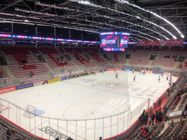 Ein Eishockeyspiel in einer großen Arena, mit Zuschauern auf Sitzen und Stehen, einem Zaun um die Eisbahn, einem Display mit der Aufschrift "St. Louis Blues vs Detroit Red Wings", Metallrahmen und einer Decke mit Deckenleuchten.