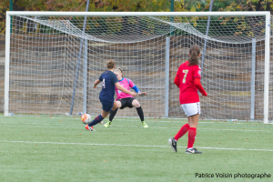 Eine Gruppe von Frauen, die auf einem Rasenfeld Fußball spielen, umgeben von Bäumen, mit einem Tor im Hintergrund und Text am unteren Rand des Bildes.