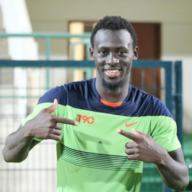 Ein Mann in einem grün-blauen Hemd, der in die Kamera lächelt und zeigt, mit einem unscharfen Geländer, Wand und Tür im Hintergrund.