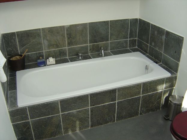 Eine weiße Badewanne mit Hähnen und verschiedenen Gegenständen in Weiß, Blau und Schwarz, ein Müllbehälter und eine Toilette auf der rechten Seite, eine geflieste Wand im Hintergrund und ein hängendes Tuch auf der linken Seite.