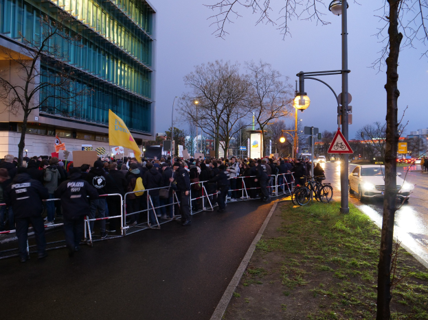 Eine große Gruppe von Menschen protestiert vor einem Gebäude in Berlin, hält Schilder und steht hinter Barrikaden, mit Fahrrädern, Laternen, Schautafeln, Bäumen und Gras im Hintergrund.