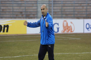 Ein Mann in einer blauen Jacke steht auf einem Fußballfeld, umgeben von Tafeln mit Text und Logos, und sieht entschlossen aus.