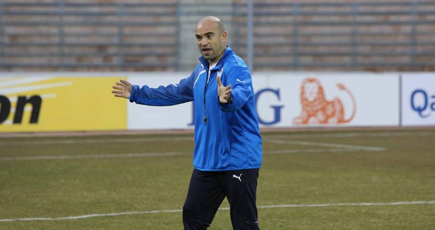 Ein Mann in einer blauen Jacke steht auf einem Fußballfeld, umgeben von Tafeln mit Text und Logos, und sieht entschlossen aus.