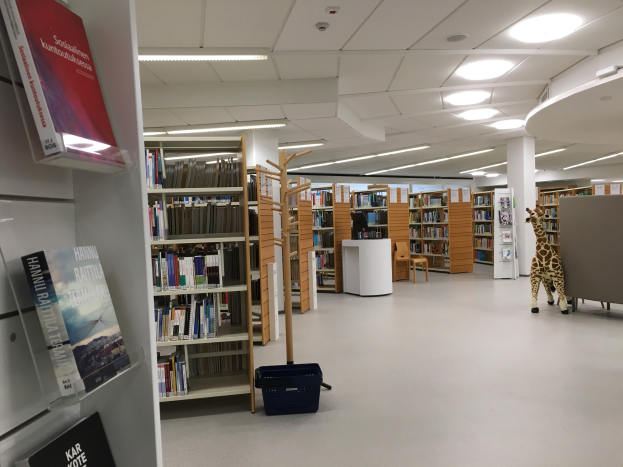 Ein geräumiger Bibliotheksinnenraum mit zahlreichen Bücherregalen, die mit Büchern gefüllt sind, ein Korb auf dem Boden, ein Giraffen-Spielzeug, Stühle, ein Tisch, Säulen und Deckenleuchten.