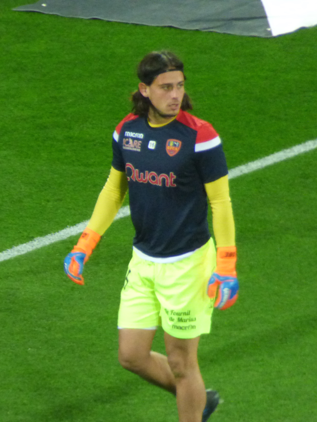 Ein Mann in einem blauen Hemd und gelben Shorts läuft auf einem Fußballfeld und streckt die Arme aus.