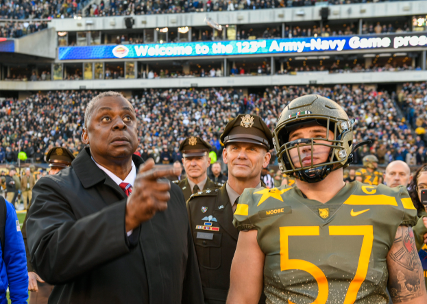 Ein Mann in Anzug und Krawatte steht neben einem Football-Spieler in einem Helm, umgeben von Menschen in Uniformen und Mützen, mit einem Stadion voller Zuschauer und einem Schild im Hintergrund, das "Oregon Football Coach Mike McCarthy und Oregon QB Marcus Mariota" anzeigt.
