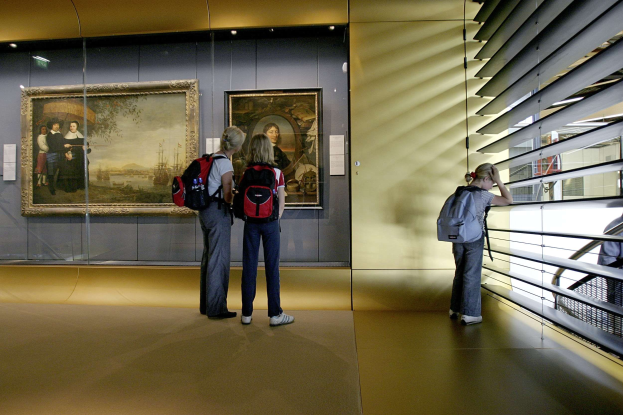 Drei Personen in einem Museum betrachten Gemälde an der Wand, mit einem Geländer auf der rechten Seite und warmem Deckenlicht.