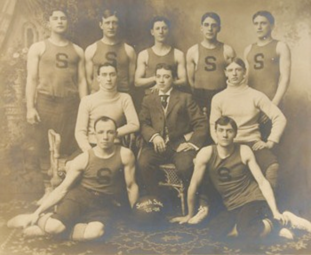 Ein Schwarz-Weiß-Foto einer Basketballmannschaft, mit Spielern, die sitzen und stehen, einer Person in der Mitte auf einem Stuhl und einem Ball vor ihnen auf dem Boden, vor einer Wand.