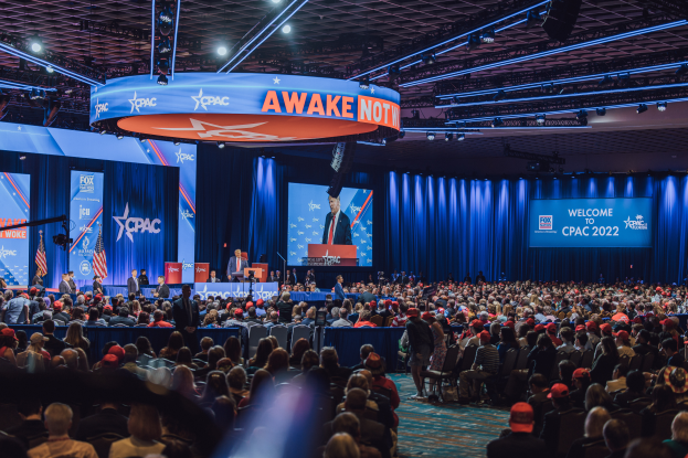 Eine große Menschenmenge, die vor einer Bühne auf einem CPAC 2020-Event sitzt, mit ein paar Personen auf dem Podium, blauen Vorhängen und Schildern im Hintergrund sowie einer Decke mit Lampen oben.