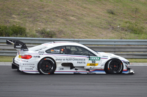 BMW M4 DTM Rennauto auf einer Rennstrecke mit Sponsorentext, umgeben von grünem Gras und einer Begrenzung im Hintergrund.