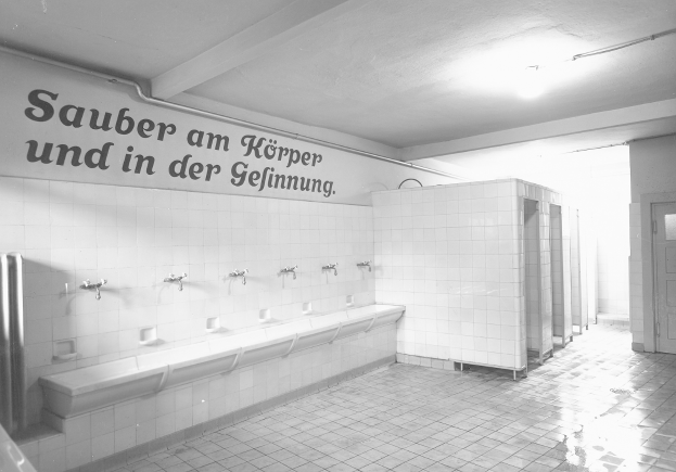 Ein Schwarz-Weiß-Foto einer öffentlichen Toilette mit einem gefliesten Boden, einer textbedeckten Wand, mehreren Hängen, einer Tür und einer Deckenlampe.