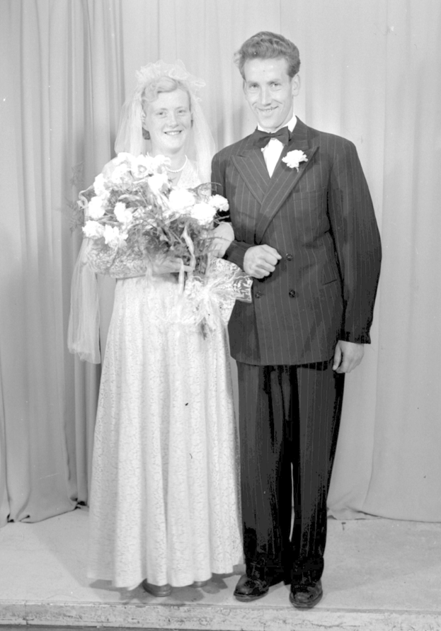 Ein Mann und eine Frau stehen nebeneinander auf einer Bühne, lächeln und die Frau hält einen Blumenstrauß in der Hand, und ein Vorhang ist hinter ihnen zu sehen.