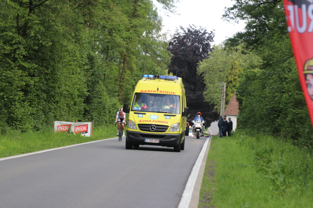 Ambulanz fährt auf einer Straße mit Fahrradfahrern nebenher, Gras und Bäume auf beiden Seiten, Häuser, Pfosten und einen klaren blauen Himmel im Hintergrund.