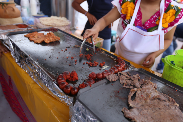 Eine Frau in einer Schürze kocht auf einem Grill an einem Markt, mit einem Tisch in der Nähe, auf dem Tabletts mit Lebensmitteln, ein Eimer und andere Gegenstände stehen, während im Hintergrund einige Menschen stehen.