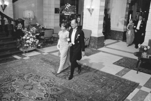 Ein Brautpaar geht durch die Hotellobby, umgeben von Menschen, Möbeln und Pflanzen, auf einem Schwarz-Weiß-Bild.