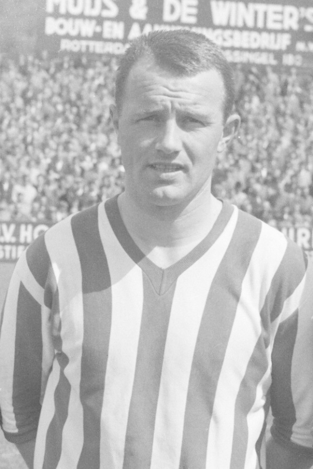 Ein Schwarz-Weiß-Foto eines Mannes in einem gestreiften Hemd, der vor einer Menge mit einer Tafel im Hintergrund steht, der mutmaßlich der legendäre niederländische Fußballspieler Bertrand ist.