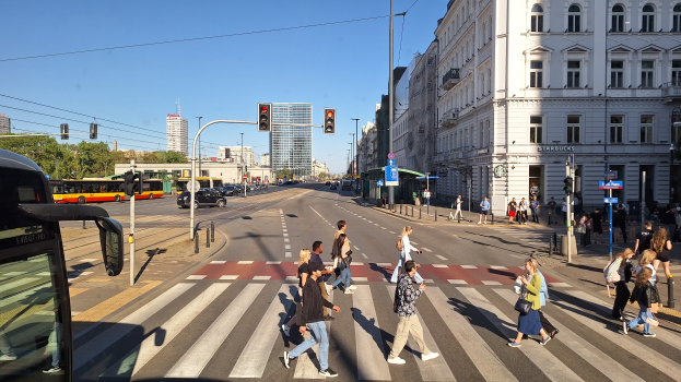 Menschen überqueren eine Straße an einer Ampel mit einem Bus, Fahrzeugen, Verkehrszeichen, Laternen, Gebäuden, Bäumen und einem klaren Himmel.