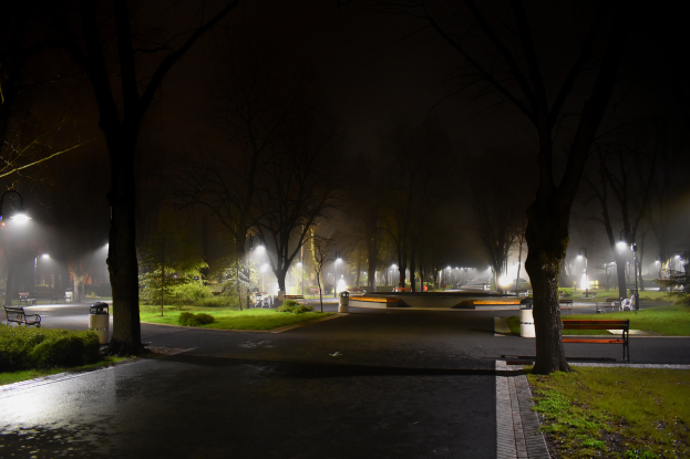 Ein Park bei Nacht mit beleuchteten Wegen, Bänken, Gras, Pflanzen und Bäumen unter einem sichtbaren Himmel mit einem Wasserzeichen auf dem Bild.