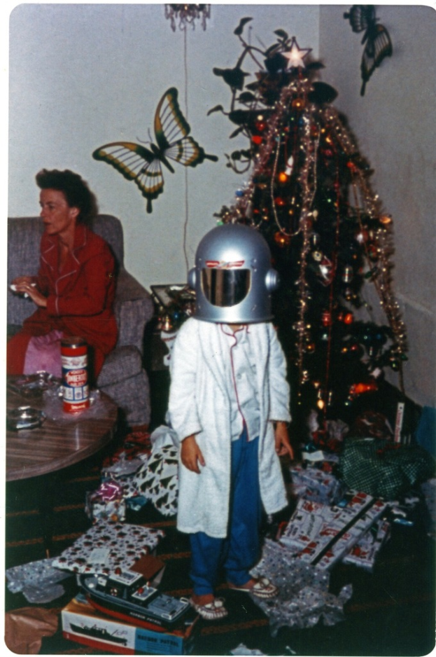 Ein Kind in einem Helm steht auf einem Boden umgeben von Geschenkverpackungen, Schachteln und einem Weihnachtsbaum, während eine Frau in einem roten Kleid auf einem Sofa sitzt mit einer Flasche und anderen Gegenständen auf einem nahen Tisch, mit zwei Schmetterlingsaufklebern auf der weißen Wand im Hintergrund.