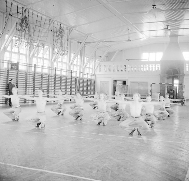 Schwarzes und weißes Bild von Männern, die Gymnastik machen und die Arme ausstrecken, in einem Turnsaal umgeben von Fenstern, einem Geländer und Deckenlampen.