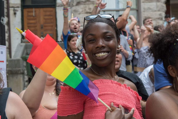 Eine Gruppe von Menschen steht vor einem Gebäude, wobei eine Frau in der Mitte eine Regenbogenflagge hält; das Gebäude hat Fenster und eine Tür und ein Schild mit Text ist sichtbar.