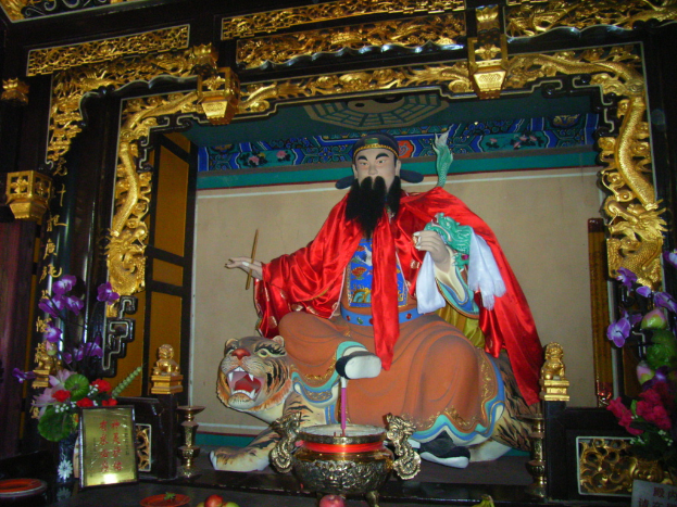 Eine Statue eines chinesischen Mönchs in buntem Gewand sitzt auf einem Tiger, der sich in einem Bogen mit goldenen Verzierungen befindet, vor einem Blumenstrauß und einer Schale in der Mitte.