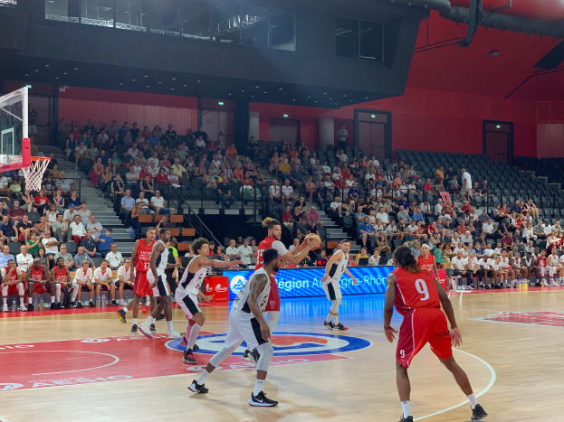 Eine Gruppe von Männern, die Basketball in einer Turnhalle mit einem Netz auf der linken Seite spielen, Zuschauer sitzen im Hintergrund und Stadionlampen darüber.