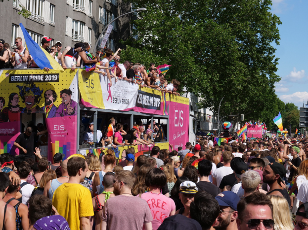 Eine große Menschenmenge marschiert auf einer Straße in Berlin Pride 2019, viele tragen Mützen und Schutzbrillen, halten Fahnen, mit Bannern und Gebäuden im Hintergrund unter einem bewölkten Himmel.