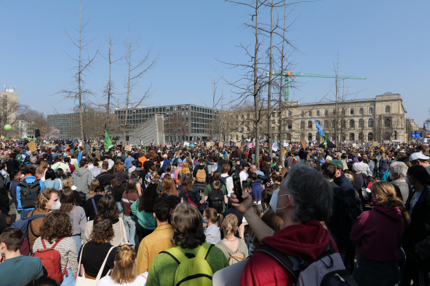 Eine große Menschenmenge mit Schildern und Taschen steht vor einem Gebäude mit Fenstern, Bäumen und einem klaren Himmel im Hintergrund, was auf eine Klimaprotest Demonstration hinweist.