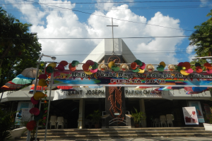 Gebäude mit einer "Frohen Festtagsgrüße St. Josef der Arbeiter"-Schrifttafel, Treppe zum Eingang, ein Bogen mit hängenden Dekorationen, Außensitzbereiche mit Tischen und Stühlen, Topfpflanzen, Bäume auf beiden Seiten und ein bewölkter Himmel.