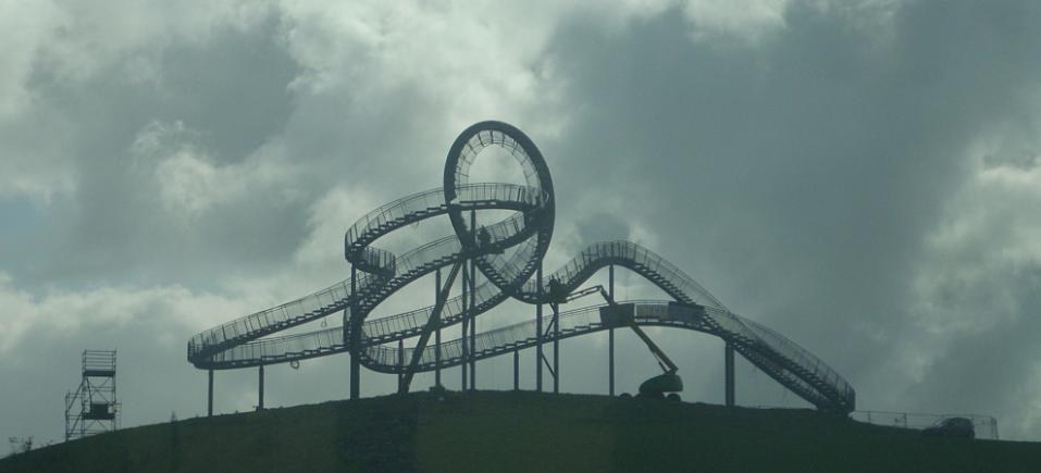 Eine Achterbahn auf dem Höhepunkt eines Hügels unter einem bewölkten Himmel, umgeben von Stützpfählen und Fahrgeschäftsinfrastruktur.