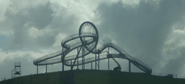 Eine Achterbahn auf dem Höhepunkt eines Hügels unter einem bewölkten Himmel, umgeben von Stützpfählen und Fahrgeschäftsinfrastruktur.