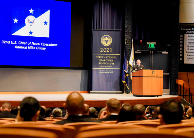 Admiral Mike Gilday steht an einem Podium mit einem Mikrofon und einer Tafel, spricht zu einer sitzenden Menge auf der 2021 Internationalen Seemacht-Symposium, mit Flaggen, einem Bildschirm mit Text und einer Treppe mit Geländern im Hintergrund.