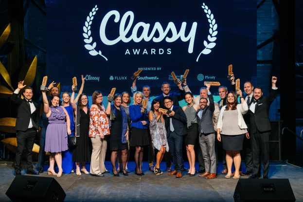 Eine Gruppe von Menschen auf einer Bühne, die strahlen und Preise halten, mit Lautsprechern unten und einem Hintergrundbild mit der Aufschrift "Classy Awards 2019".