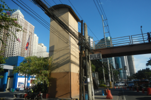 Eine Stadtstraße mit hohen Gebäuden, Fahrzeugen, Verkehrskegeln, Strommasten mit Kabeln, einer Brücke mit Geländern, einem Namensschild, Bäumen und einem bewölkten Himmel.