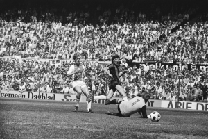 Schwarzes Foto von Männern, die auf einem Feld Füssball spielen, mit Zuschauern im Hintergrund und einem Banner mit der Aufschrift "Bundesliga 1978-79 Ajax 1-1 Hannover 96".