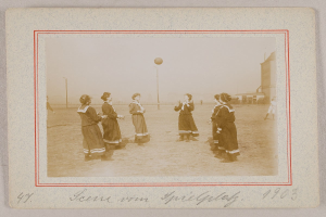 Eine Gruppe von Frauen, die auf dem Rasen Fußball spielen, mit einem Ball in der Mitte, Pfosten und Gebäuden im Hintergrund und Text am unteren Bildrand.