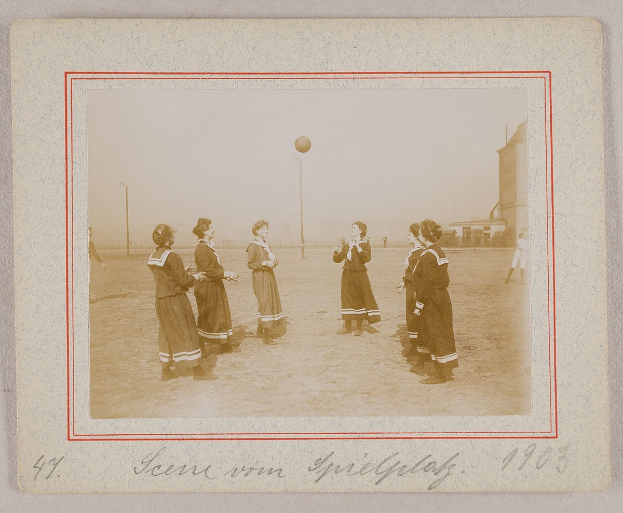 Eine Gruppe von Frauen, die auf dem Rasen Fußball spielen, mit einem Ball in der Mitte, Pfosten und Gebäuden im Hintergrund und Text am unteren Bildrand.