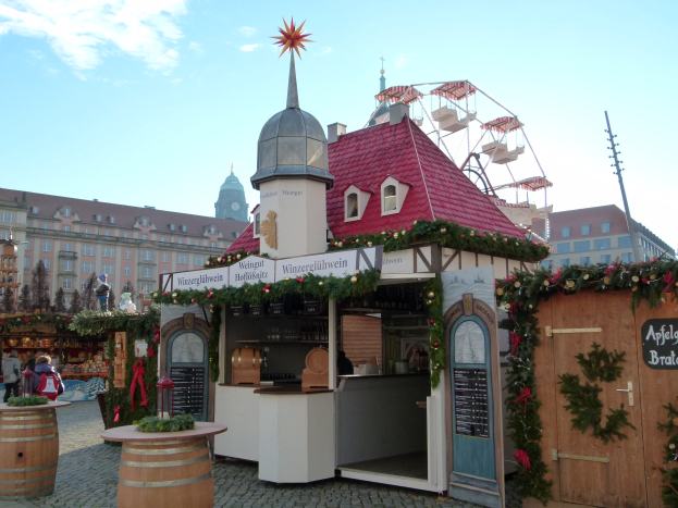 Kleines Gebäude mit rotem Dach und Riesenrad davor, umgeben von Festdekorationen und Menschen bei einem Outdoor-Event mit Gebäuden, Bäumen und bewölktem Himmel im Hintergrund.
