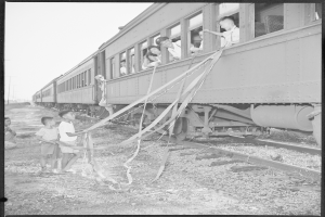 Ein Schwarz-Weiß-Foto einer Gruppe von Kindern, die neben einem Zug auf einem Bahngleis stehen, einige halten Bänder, mit Pfählen und einem klaren Himmel im Hintergrund.
