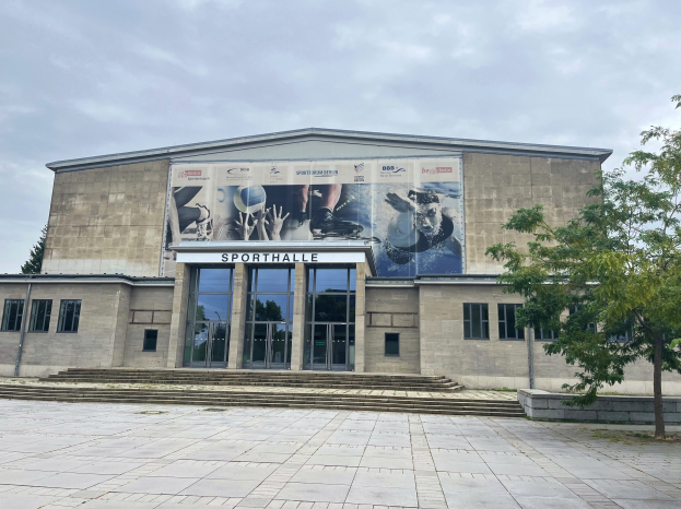 Ein großes Gebäude mit einer Wandmalerei an der Seite, umgeben von Bäumen auf der rechten Seite und einem bewölkten Himmel im Hintergrund, identifiziert als Sporthalle in Berlin, Deutschland.