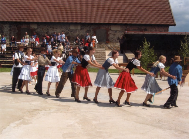 Eine Gruppe von Menschen in traditioneller bayrischer Tracht tanzt vor einem Gebäude mit Dach und Fenstern, einige tragen Hüte und halten Stöcke, andere sitzen auf Bänken, mit grasbewachsenem Boden und Bäumen im Hintergrund unter einem bewölkten Himmel.