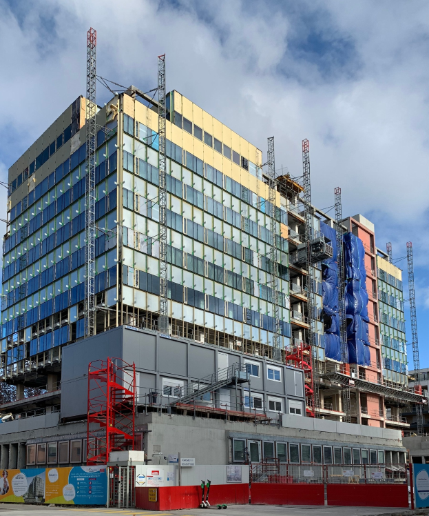 Großes Gebäude im Bau in einer städtischen Umgebung mit Glasfenstern, Metallrahmen, einer Treppe, einem Zaun, einer Tafel, Pfosten, einem Straßenpfosten, Bäumen und einem bewölkten Himmel.