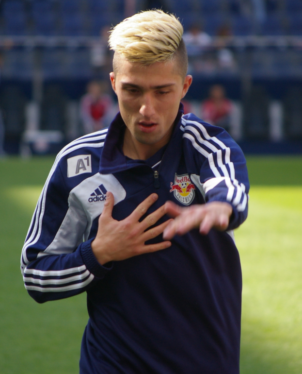 Ein junger Mann in einem blauen T-Shirt steht auf einem Fußballfeld und streckt die Arme aus, mit entschlossener Miene und unscharfem Hintergrund.