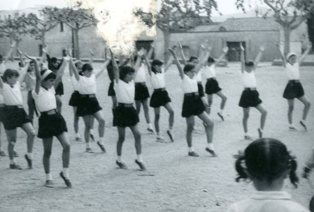 Schwarzes und weißes Foto von jungen Mädchen in Schuluniformen, die auf einer von Gebäuden und Bäumen gesäumten Straße tanzen, mit einer Person in Schuhen unten sichtbar.