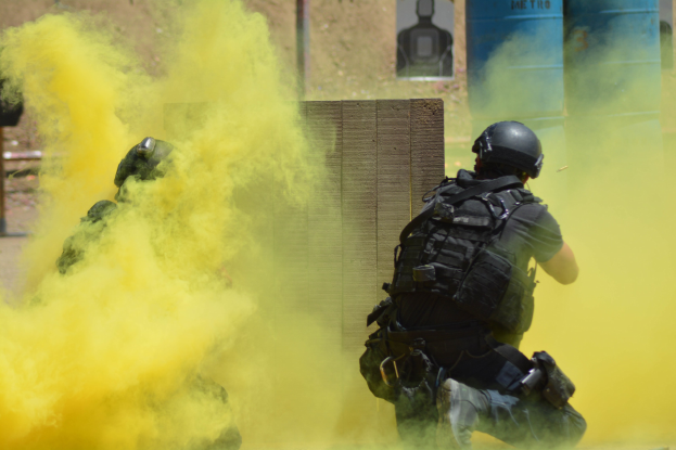 Zwei Polizisten in Einsatzausrüstung mit gelbem Rauch aus dem Mund, tragen Helme und Schutzausrüstung und stehen vor einer Wand mit einer Tafel und anderen Gegenständen, wobei der Boden unten sichtbar ist.